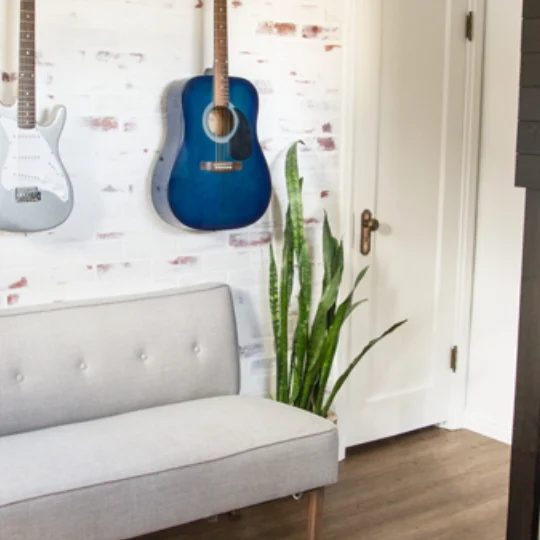 Red Brick Wall Painted White With Red Accents Coming Through Guitars Hanging On Wall, Grey Couch And Green Plant