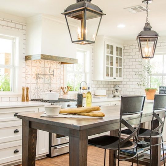 Fixer Upper Worm House Kitchen With Wood Table And White Brick Walls, White Cabinets And White Vent Hood