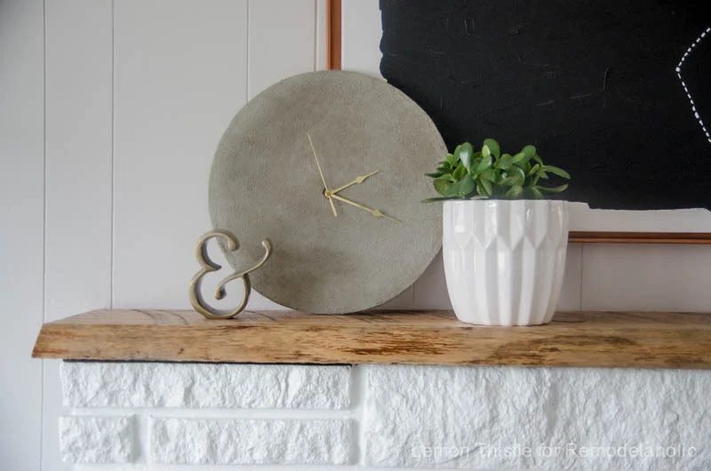 direct Handmade Concrete Clock With Gold Hands Sitting On Wooden Shelf Above White Brick Mantle And White Planter With Greenery