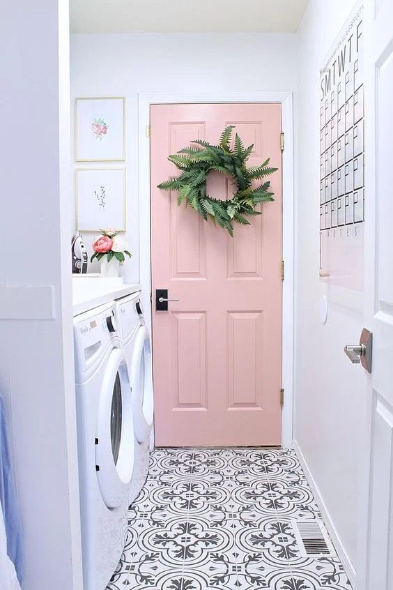 Grey And White Patterned Floor Of Long Narrow Laundry Room, White Front Load Washer And Dryer On The Side, Calendar On Opposite Wall, Pale Pink Door With Greenery Wreath At End
