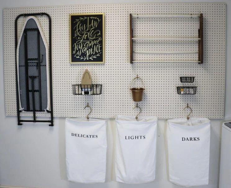 Laundry Room Pegboard With Hanging Laundry Bags Labeled Delicates, Lights, And Darks Ironing Board Also Hanging
