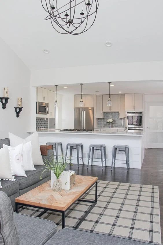 White Open Kitchen And Living Space, Metal Bar Stools, Wooden Table, Grey Couch With White Windows, Chrome Kitchen Appliances