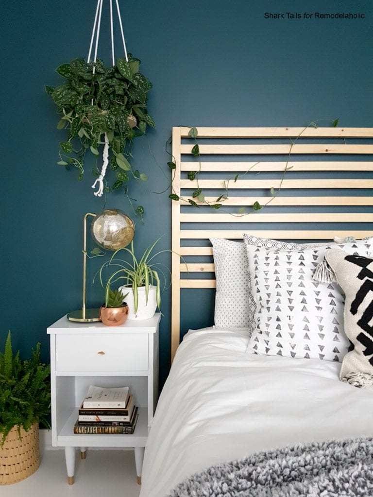 Blue Wall With Natural Wood Headboard, White Bedding, Patterned Pillows And Hanging Plant From The Ceiling, White Nighstand With Gold Accented Light And Planter